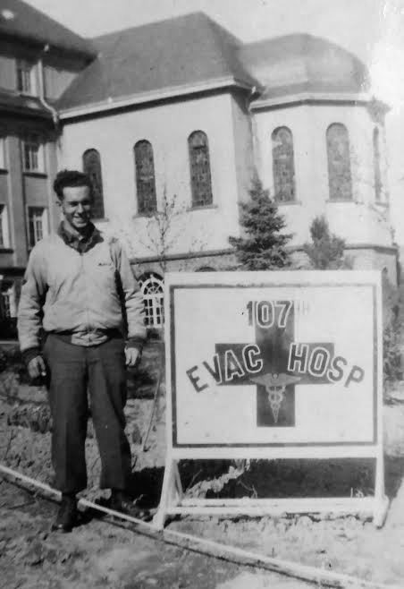 Picture illustrating Technician 5th Grade Robert W. Frisbie, with sign indicating the 107th Evacuation Hospital. Taken during the organization’s stay at the Pensionnat St. Joseph, Hachy, Belgium, 21 January 1945.