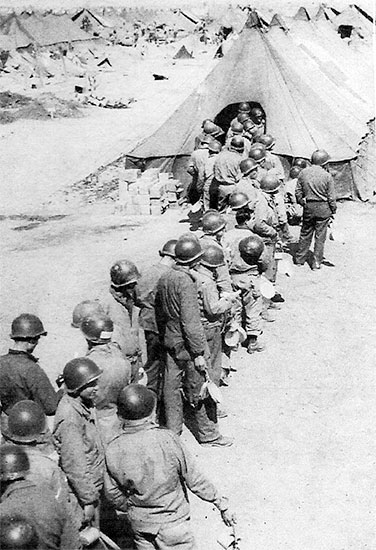 Picture illustrating the 56th Evacuation Hospital chow line for Enlisted Men. Wearing steel helmets eventually became compulsory because of the constant bombing and shelling by enemy long-range artillery and aircraft.